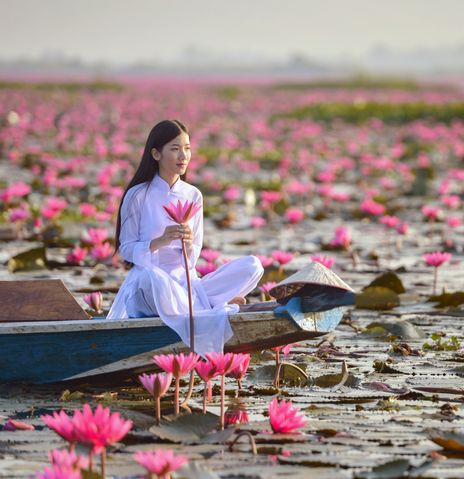 Frau im weißen Kleid sitzt in kleinem Boot auf See voller rosa Seerosenblüten