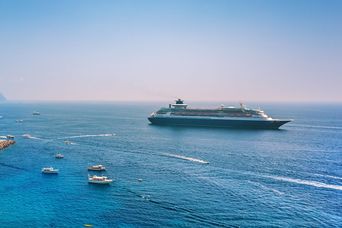 Ein Kreuzfahrtschiff liegt vor einer Küste im ruhigen blauen Meer, kleinere Boote kreuzen die Bucht.