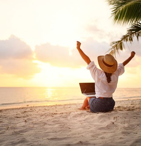 Frau sitzt mit Laptop am Strand vor dem Meer und genießt den Sonnenaufgang