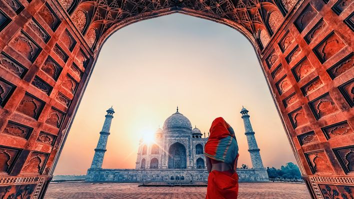 Frau im roten Sari blickt durch einen Torbogen auf das Taj Mahal in Agra, Indien