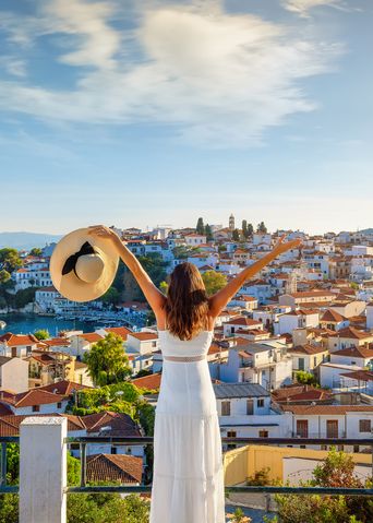 Eine Frau im weißen Kleid mit einem Hut in der Hand steht auf einer Aussichtsplattform und schaut in die Stadt Skiathos in Griechenland