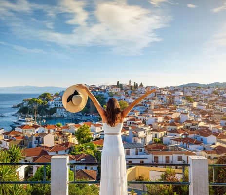 Eine Frau im weißen Kleid mit einem Hut in der Hand steht auf einer Aussichtsplattform und schaut in die Stadt Skiathos in Griechenland