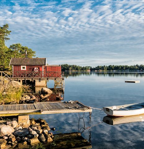 Ein rotes Holzhaus und ein kleines Boot liegen an einem ruhigen, bewaldeten See in der Natur Schwedens.