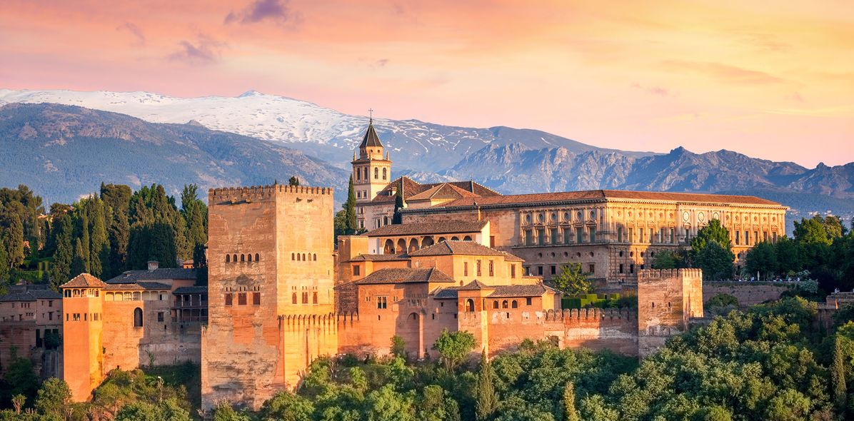 Die Alhambra leuchtet bei Sonnenuntergang vor der Sierra Nevada in warmen Farben über Granada.