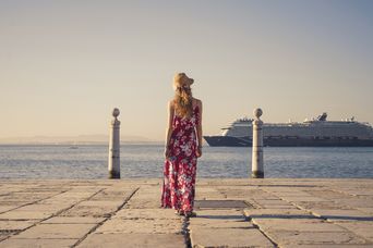 Frau mit einem bunten langen Kleid steht an einer Promenade und schaut auf ein Kreuzfahrtschiff