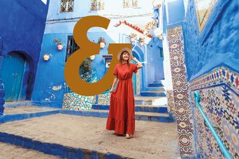 Eine Frau im Roten Kleid mit Hut spaziert durch die blauen Gassen von Chefchaouen und bestaunt die Architekttur mit dem Berge & Meer Superzeichen