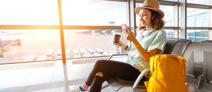 Frau mit Hut sitzt in einem Flughafen-Terminal am Fenster zum Rollfeld mit Handy und Kaffeebecher neben einem gelben Rucksack
