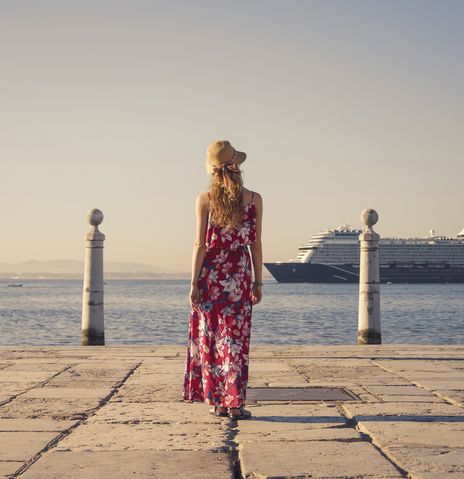 Frau mit einem bunten langen Kleid steht an einer Promenade und schaut auf ein Kreuzfahrtschiff