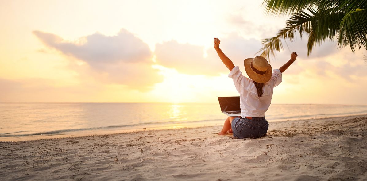Eine Frau sitzt im Sand und auf dem Schoß ein Laptop