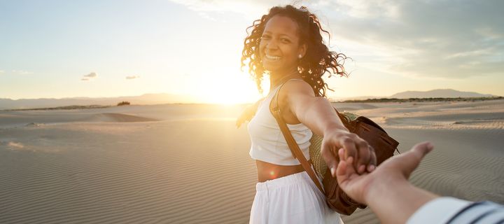 Frau im weißen Kleid dreht sich lachend am Strand und hält die Hand ihres Partners