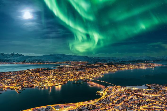 Blick auf die verschneite Stadt Tromso von oben bei Nacht und Nordlichter am Himmel, Norwegen