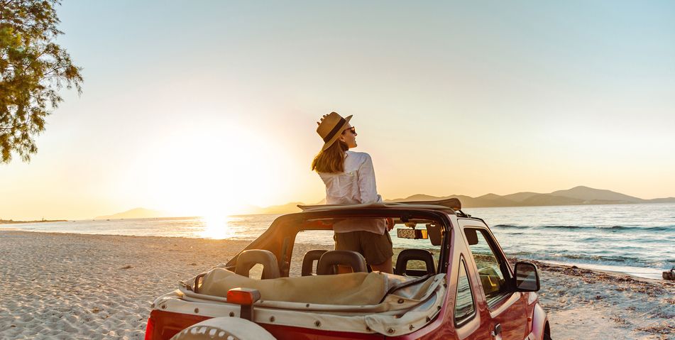 Eine Frau mit Ihrem Auto steht an einem Strand und beobachtet den Sonnenuntergang über dem Meer