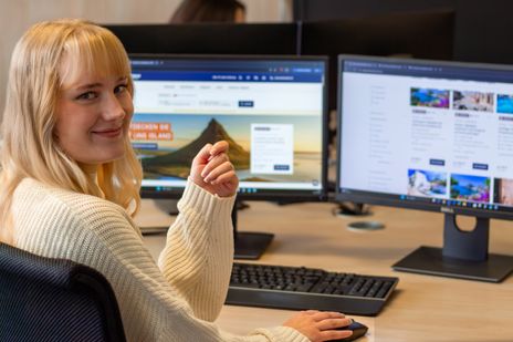 Auszubildende Denise sitzt an ihrem Arbeitsplatz und lächelt in die Kamera