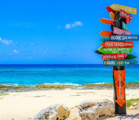 Ein bunter Wegweiser mit Ländern der Welt steht auf eine weißen Sandstrand vor blauem Wasser und blauem Himmel