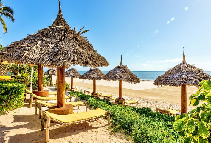 Blick aufs Meer von Sansibar über einen typischen Strand mit Sonnenschirmen und -liegen aus Palmen und Bäumen