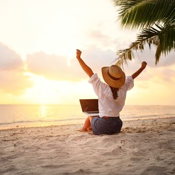 Frau sitzt im Sand und hat einen Laptop auf dem Schoß