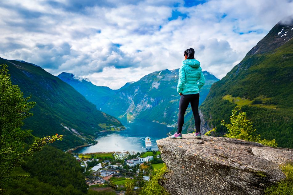 Eine Frau steht auf einem Felsvorsprung und schaut in einen Fjord hinunter wo ein Schiff zu sehen ist